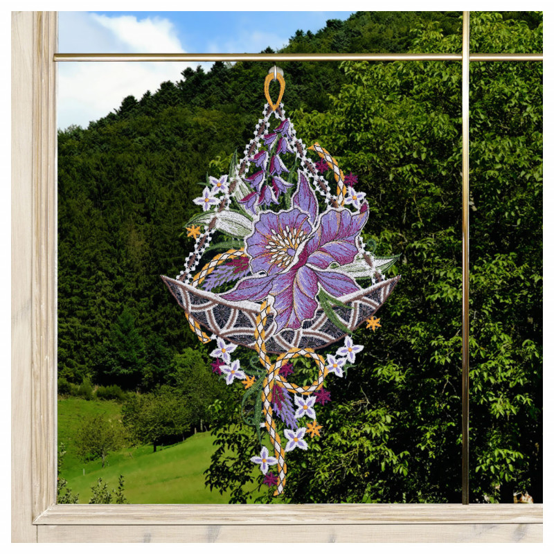 Ein Spitzenornament mit einer auffälligen violetten Blüte und verschlungenen Mustern hängt in einem Fenster. Durch das Glas ist ein dicht mit grünen Bäumen bewachsener Hügel unter einem klaren Himmel zu sehen, der einen lebendigen natürlichen Hintergrund für die Dekoration bildet.