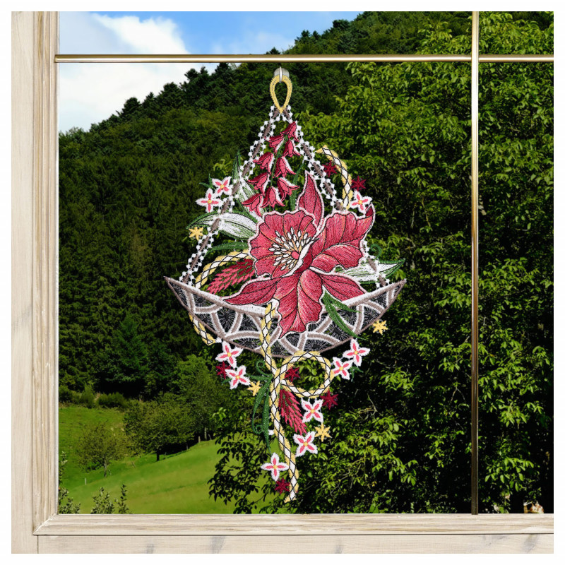 Ein gesticktes Blumenornament mit einer auffälligen roten Blüte, grünen Blättern und kleinen rosa Details hängt in einem Fenster. Außerhalb des Fensters befinden sich grüne Bäume und ein Hügel, die eine natürliche Hintergrundszene bilden.