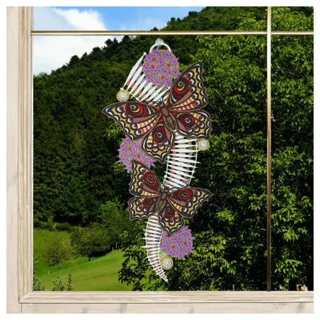 Ein dekorativer Aufhänger zeigt zwei schwarze, rote und weiße Schmetterlinge mit detaillierten Mustern, die auf violetten Stachelblumen sitzen. Das Stück ist in einem Fenster ausgestellt, wobei durch das Glas im Hintergrund grüne Bäume und Hügel zu sehen sind.