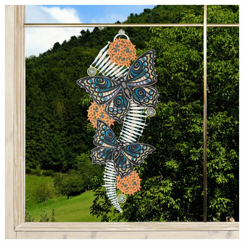 Ein Fensterbehang zeigt zwei blaue und schwarze Schmetterlinge mit verschnörkelten Mustern, begleitet von orangefarbenen Blumendetails und weißen geschwungenen Mustern. Durch den Holzrahmen des Fensters ist im Hintergrund eine grüne Waldlandschaft zu sehen.