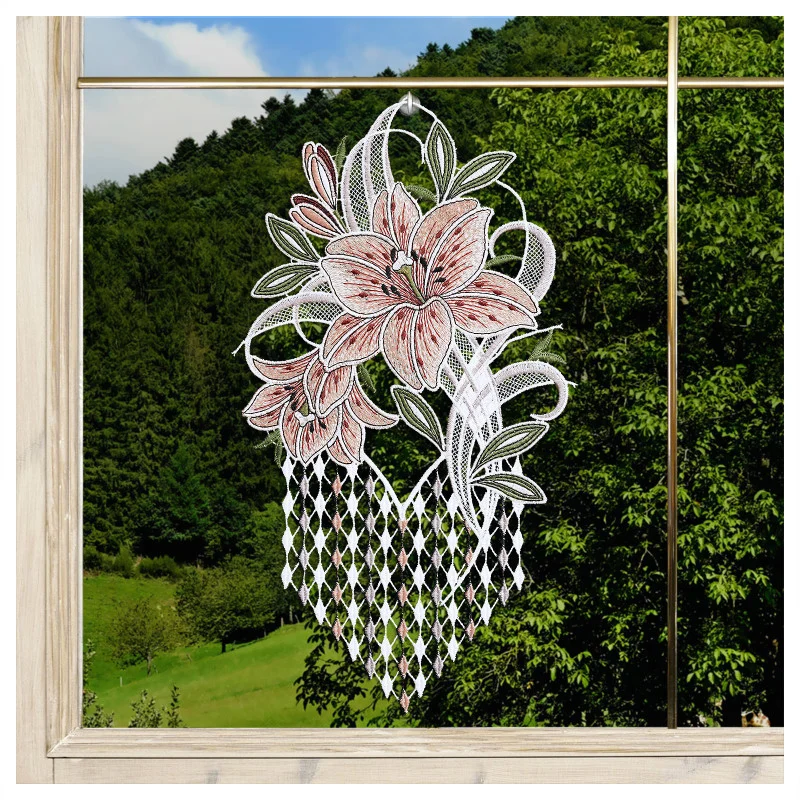 Eine Fensterdekoration aus Spitze mit rosa und weißen Blumen und grünen Blättern ist auf einer Fensterscheibe abgebildet. Das Fenster hat einen hellen Holzrahmen. Durch das Glas sind sanfte grüne Hügel, Bäume und ein blauer Himmel mit vereinzelten Wolken zu sehen.
