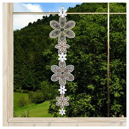 Eine vertikale Kette aus detaillierten, weißen, spitzenartigen Blumenornamenten ist vor einem Fenster aufgehängt. Außerhalb des Fensters befinden sich grüne Bäume, grasbewachsene Hügel und ein bewaldeter Berg unter einem teilweise bewölkten blauen Himmel. Der Fensterrahmen ist aus Holz gefertigt.