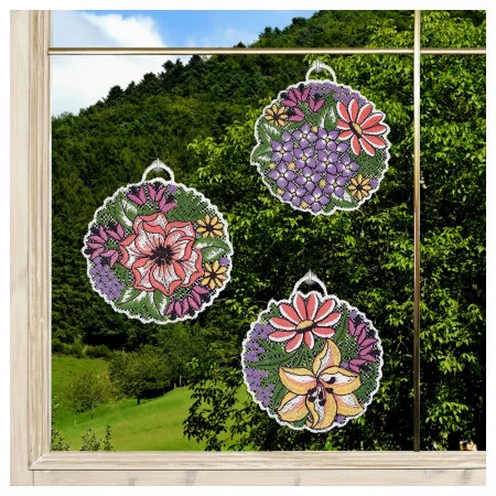 Drei runde Sonnenfänger mit floralen Mustern und farbenfrohen Blumenmotiven hängen an einem Fenster. Hinter dem Glas sind grüne Hügel und Bäume vor einem blauen Himmel zu sehen. Das Fenster ist mit Holz umrahmt.