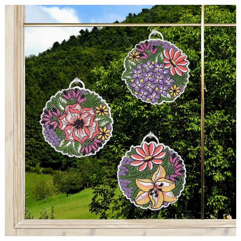 Drei runde Sonnenfänger mit floralen Mustern und farbenfrohen Blumenmotiven hängen an einem Fenster. Hinter dem Glas sind grüne Hügel und Bäume vor einem blauen Himmel zu sehen. Das Fenster ist mit Holz umrahmt.