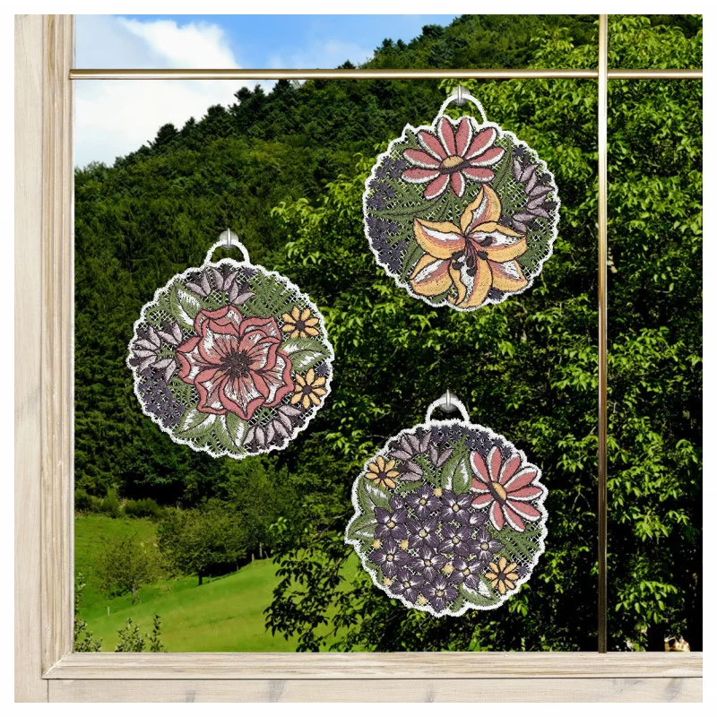 Drei runde Sonnenfänger mit Blumenmustern hängen an einem Fenster. Jeder Sonnenfänger zeigt detaillierte, bunte Blumen und grüne Blätter. Außerhalb des Fensters befinden sich grüne Bäume und grasbewachsene Hügel unter einem klaren Himmel, der von der hölzernen Fensterstruktur eingerahmt wird.