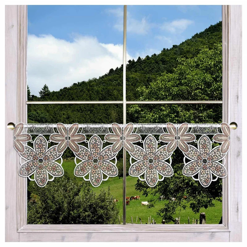 Ein Fenster mit einem hellen Holzrahmen ist mit einem Spitzenvolant mit großen, detaillierten Blumenmustern in Braun und Weiß versehen. Durch das Fenster ist eine grüne Landschaft mit Bäumen, Hügeln und grasenden Kühen unter einem teilweise bewölkten Himmel zu sehen.