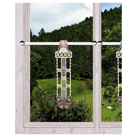 Ein Fenster mit hellem Holzrahmen bietet einen Blick auf einen grünen Hügel und Bäume. An dem Fenster hängt eine dekorative Spitzenblende mit einem großen Schmetterlingsmotiv am unteren Rand sowie floralen und geometrischen Mustern in Weiß, Orange, Rot und Gelb.