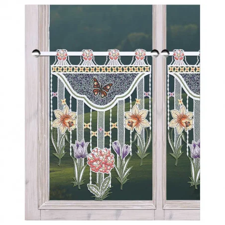 Gardinenpaneel aus Spitze mit mehrfarbiger Blumenstickerei, die Lilien, Krokusse, eine rosa Kugelprimel und einen braunen Schmetterling darstellt. Das Paneel hat gebördelte Ränder und wird oben mit Schlaufen an einer Stange aufgehängt, die ein Fenster mit Blick auf grünes Laub einrahmt.