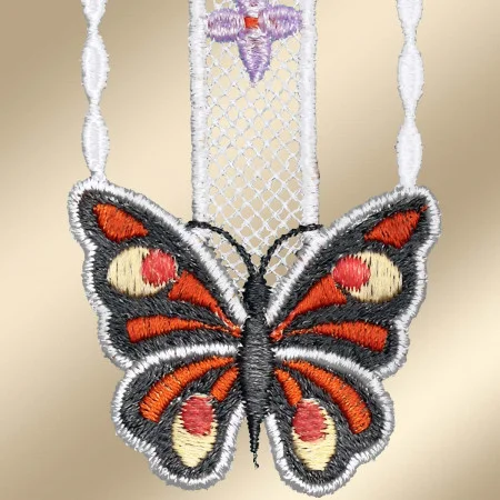 Nahaufnahme eines gestickten Schmetterlingsanhängers aus schwarzem, rotem, cremefarbenem und orangefarbenem Garn. Der Anhänger hängt an einer feinen weißen Spitzenkette mit einem kleinen lila gestickten Motiv über dem Schmetterling. Der goldene Hintergrund ist sanft verschwommen.
