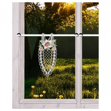 Ein Spitzenornament mit Blumenstickerei hängt an einer Stange im Inneren eines holzgerahmten Fensters. Außerhalb des Fensters fällt das Sonnenlicht durch die Äste der Bäume auf eine grüne Wiese mit gelben Pusteblumen. Das Bild ist gut beleuchtet und klar.