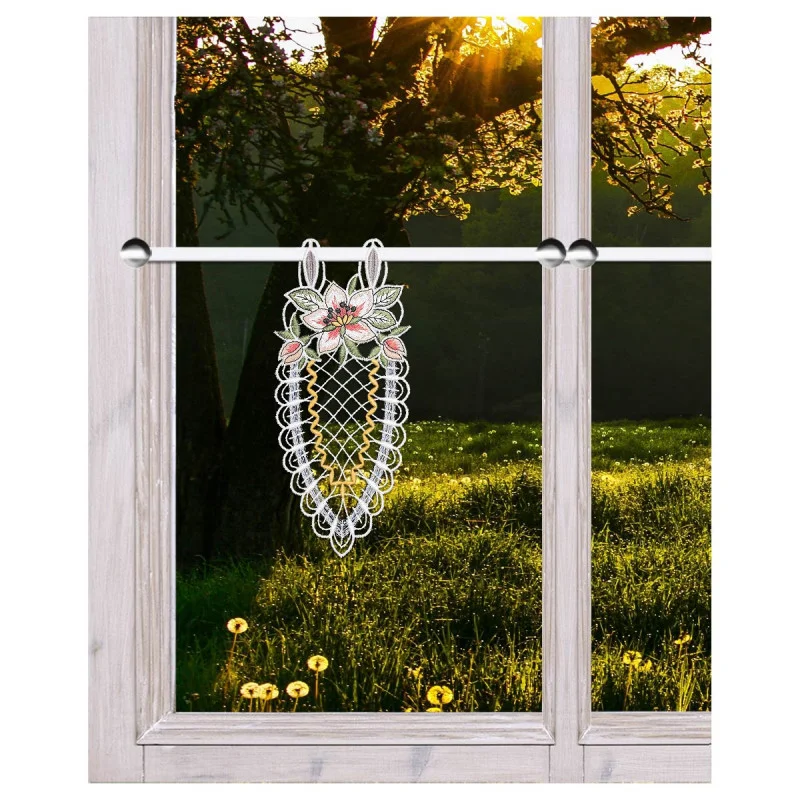 Ein Spitzenornament mit Blumenstickerei hängt an einer Stange im Inneren eines holzgerahmten Fensters. Außerhalb des Fensters fällt das Sonnenlicht durch die Äste der Bäume auf eine grüne Wiese mit gelben Pusteblumen. Das Bild ist gut beleuchtet und klar.
