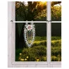 Ein Spitzenornament mit Blumenstickerei hängt an einer Stange im Inneren eines holzgerahmten Fensters. Außerhalb des Fensters fällt das Sonnenlicht durch die Äste der Bäume auf eine grüne Wiese mit gelben Pusteblumen. Das Bild ist gut beleuchtet und klar.
