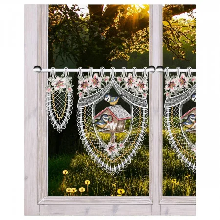 Nahaufnahme eines Fensterrahmens aus hellem Holz mit Spitzenvorhängen, die mit Vogelhäusern, Vögeln und rosa Blumen bestickt sind. Draußen fällt das Sonnenlicht durch die Bäume auf eine Wiese mit gelbem Löwenzahn.