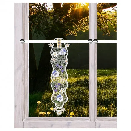 Ein Spitzenvorhang mit einem Blumenmuster in Lila, Gelb und Weiß hängt an einer Stange vor einem Fenster mit Holzrahmen. Außerhalb des Fensters fällt das Sonnenlicht durch hohe Bäume auf eine mit Löwenzahn und Wildblumen übersäte Wiese.