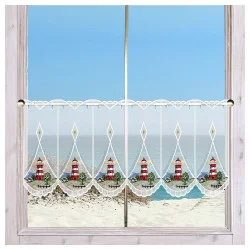 An einem Fenster mit hellem Holzrahmen hängt ein Spitzenvorhang mit rot-weißen Leuchtturm-Motiven. Der Vorhang hat gewellte Ränder und eine Zierborte. Durch das Fenster sind ein Sandstrand, grasbewachsene Dünen und ein ruhiges blaues Meer unter einem klaren Himmel zu sehen.