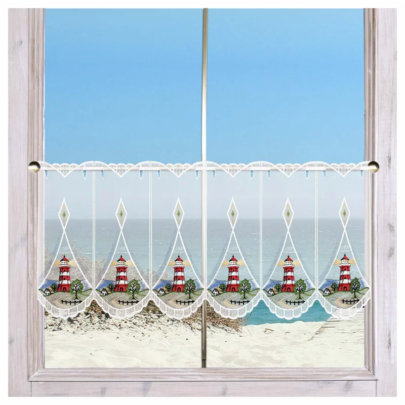 An einem Fenster mit hellem Holzrahmen hängt ein Spitzenvorhang mit rot-weißen Leuchtturm-Motiven. Der Vorhang hat gewellte Ränder und eine Zierborte. Durch das Fenster sind ein Sandstrand, grasbewachsene Dünen und ein ruhiges blaues Meer unter einem klaren Himmel zu sehen.