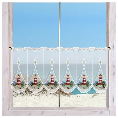 An einem Fenster mit hellem Holzrahmen hängt ein Spitzenvorhang mit rot-weißen Leuchtturm-Motiven. Der Vorhang hat gewellte Ränder und eine Zierborte. Durch das Fenster sind ein Sandstrand, grasbewachsene Dünen und ein ruhiges blaues Meer unter einem klaren Himmel zu sehen.