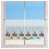 An einem Fenster mit hellem Holzrahmen hängt ein Spitzenvorhang mit rot-weißen Leuchtturm-Motiven. Der Vorhang hat gewellte Ränder und eine Zierborte. Durch das Fenster sind ein Sandstrand, grasbewachsene Dünen und ein ruhiges blaues Meer unter einem klaren Himmel zu sehen.