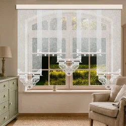 Ein Wohnzimmer mit einem beigen Sessel, einer hellgrünen Kommode und einer Tischlampe. Am Fenster hängen weiße, durchsichtige Spitzenvorhänge mit aufwendigen Mustern, die Tageslicht hereinlassen und den Blick auf das grüne Gras und die Bäume draußen freigeben.