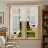 Ein Raum mit hellen Wänden verfügt über ein Fenster mit Holzrahmen, das den Blick auf grünes Gras freigibt. Transparente, weiß bestickte Vorhänge hängen in unterschiedlichen Höhen. Der Raum ist mit einem gestreiften Sessel, einem gerahmten Wandbild und einem Holzschrank ausgestattet, in dem Bücher und Dekoartikel ausge