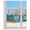 Die weiß gerahmten Fenster sind mit Spitzenvorhängen mit Schiffs- und Möwenmotiven versehen. Draußen erstrecken sich unter einem klaren Himmel ein Sandstrand, grüne Sträucher und das blaue Meer. Die Vorhänge versperren teilweise den Blick auf die Küstenlandschaft.