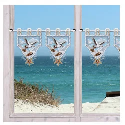 Fenster mit weißen Rahmen und transparenten Volants, die mit Vögeln und Sternen bestickt sind. Durch die Fenster sieht man grasbewachsene Sanddünen, eine hölzerne Promenade und türkisfarbene Meereswellen unter einem strahlend blauen Himmel.