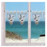 Fenster mit weißen Rahmen und transparenten Volants, die mit Vögeln und Sternen bestickt sind. Durch die Fenster sieht man grasbewachsene Sanddünen, eine hölzerne Promenade und türkisfarbene Meereswellen unter einem strahlend blauen Himmel.
