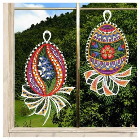 Zwei bunte Fensterbilder aus dem 2-teiligen Set "Ostereier Primel & Blütenrispe blau" (21x13 cm), gefertigt aus vogtländischer Spitze, hängen an einem Fenster. Draußen sind grüne Hügel und Bäume unter einem blauen Himmel zu sehen.